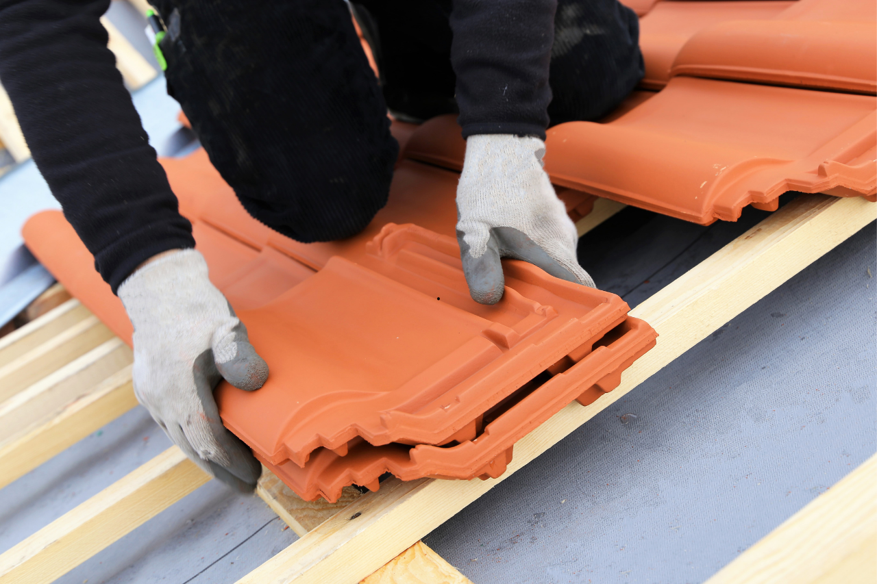 Une personne portant des gants place des tuiles de toit en terre cuite sur une charpente en bois.