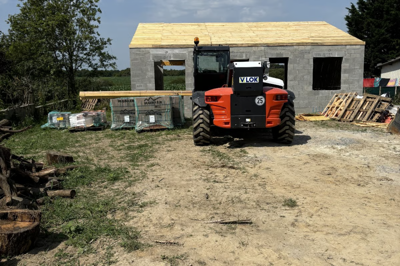 Un engin de chantier orange est stationné devant un bâtiment en béton partiellement construit, avec un toit en bois.