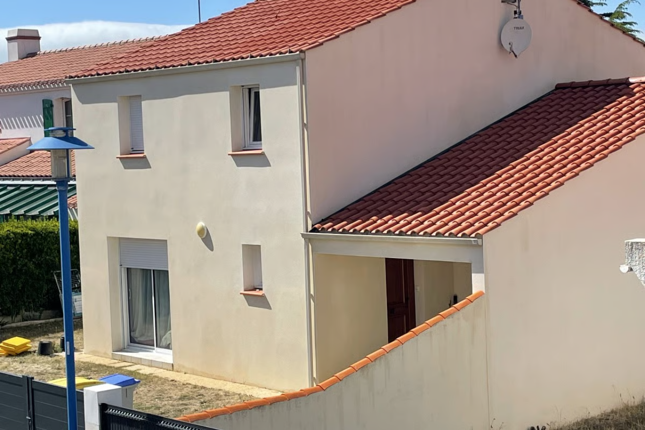 Maison à deux étages avec un toit de tuiles rouges, des murs blanc cassé et une petite véranda. Belle journée ensoleillée.