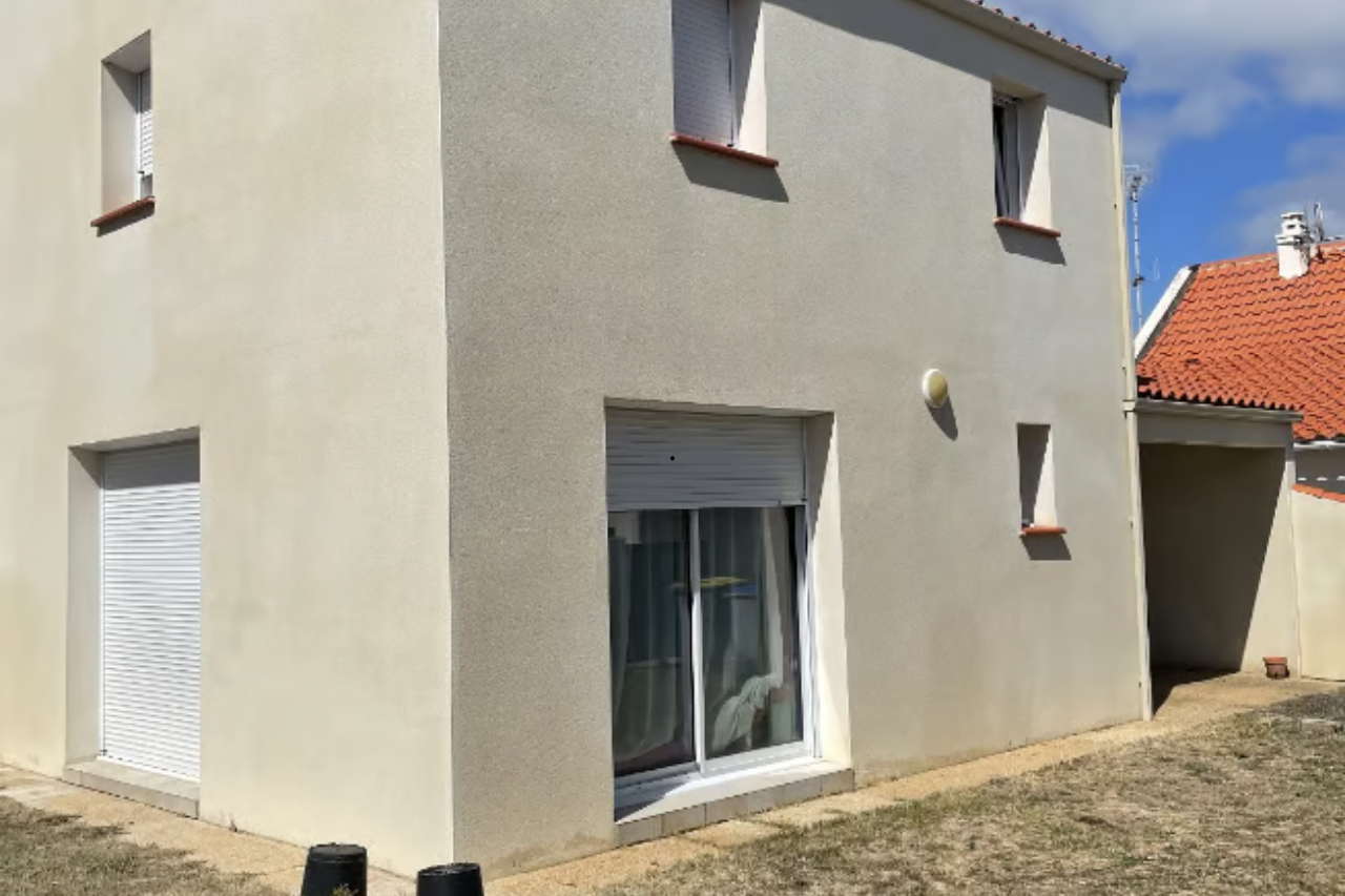 Bâtiment de deux étages avec façade en stuc beige, volets blancs et petite cour par une journée ensoleillée.