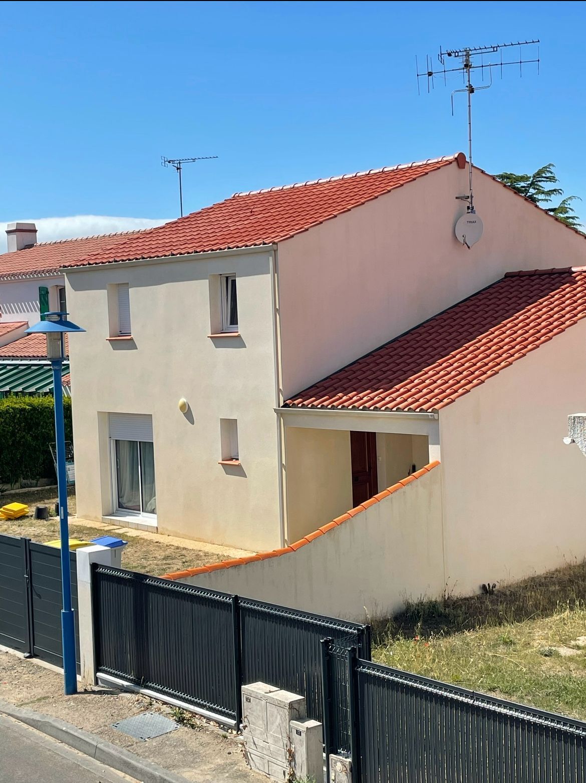 Maison à deux étages avec toit gris foncé, façade en briques claires, garage attenant, entourée de verdure et de ciel bleu.