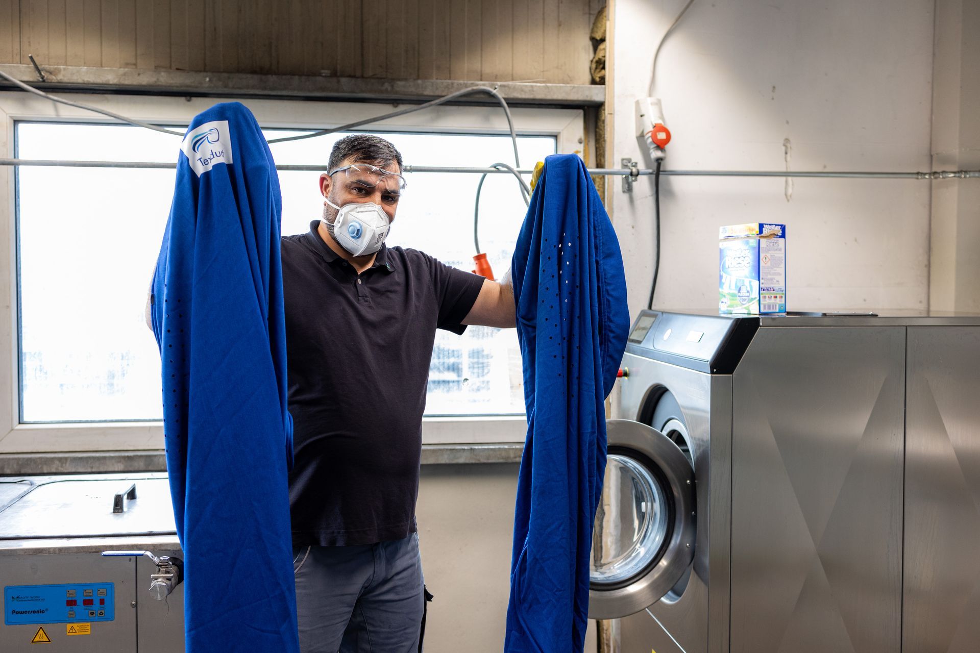 Ein Mann mit einer Maske hält zwei blaue Hemden vor einer Waschmaschine.