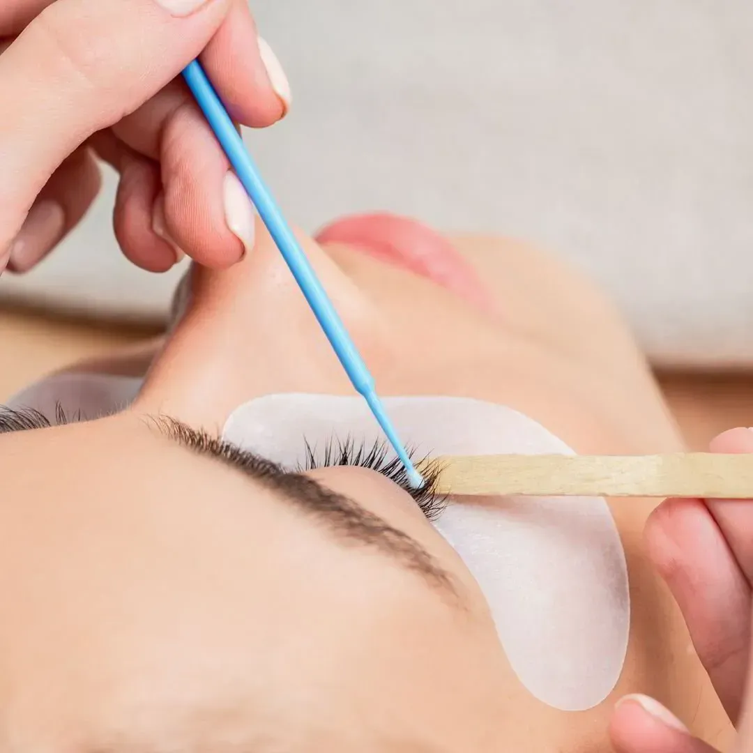 Primer plano de una esteticista aplicando extensiones de pestañas con un microaplicador azul y una herramienta de madera.