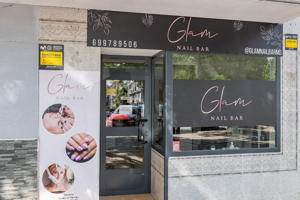 Fachada de “Glam Nail Bar” con letrero negro, puerta de cristal y un mural blanco en la pared que muestra diseños de uñas.