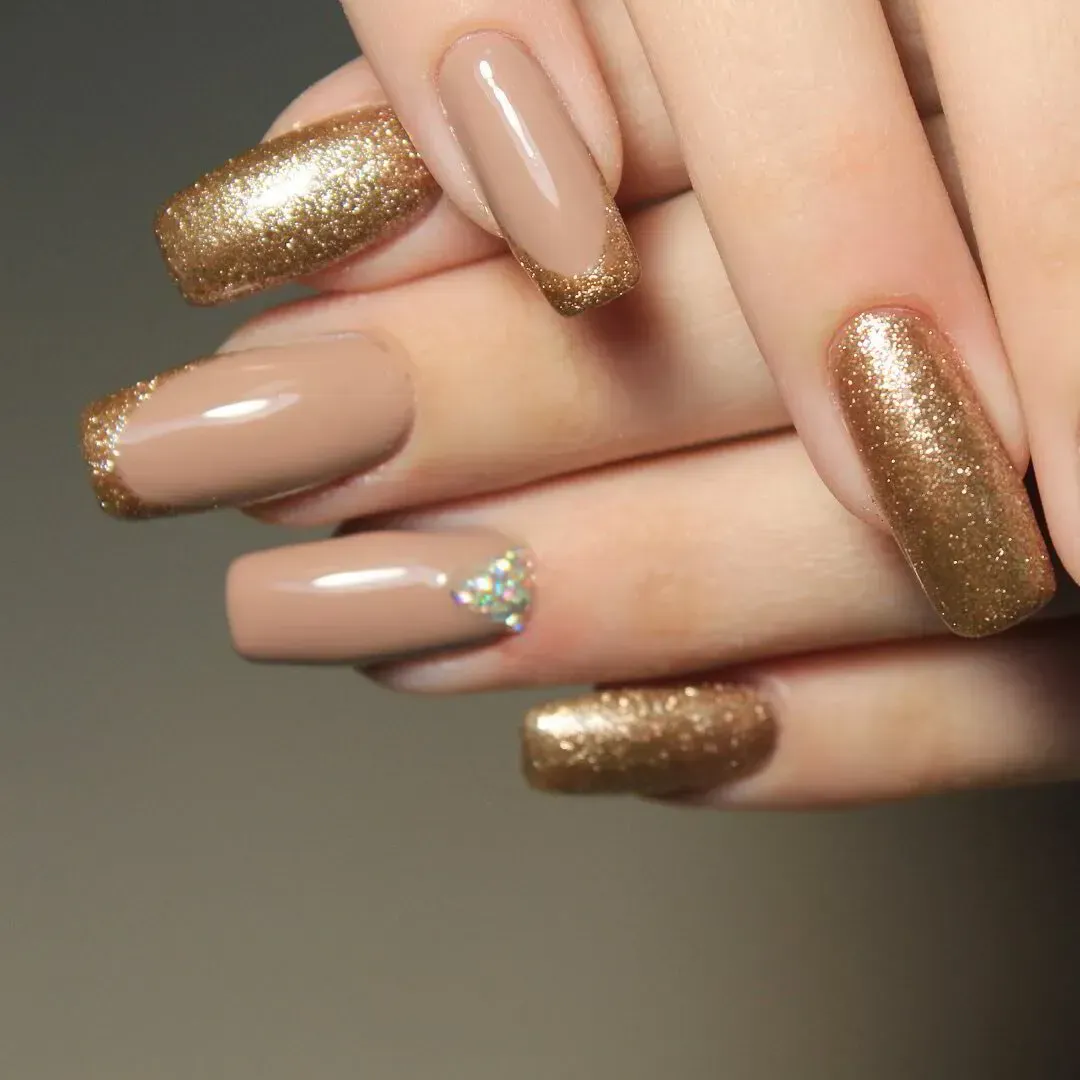 Primer plano de unas manos que muestran una manicura con esmalte nude y dorado con purpurina