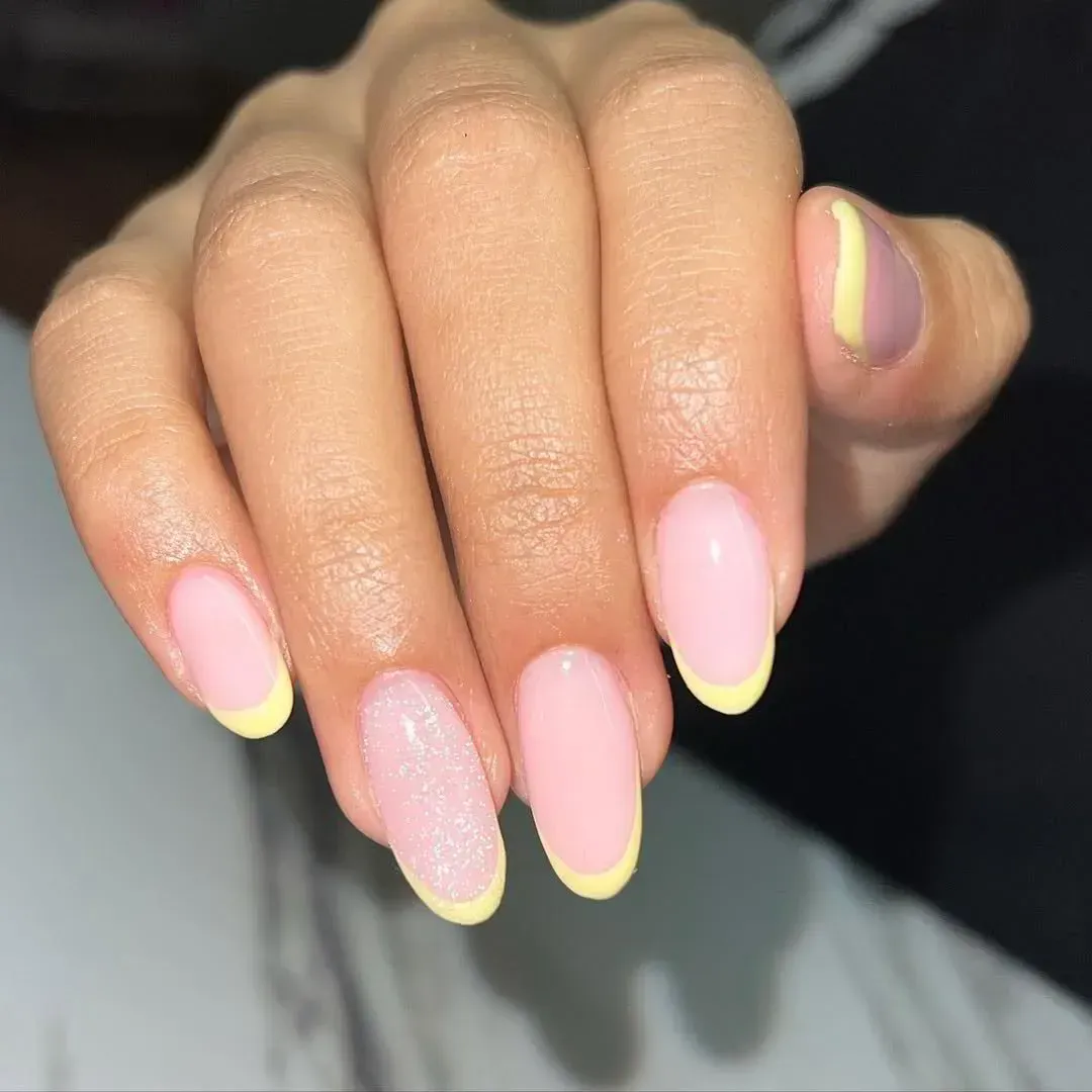 Una mano con uñas en forma de almendra, con una base rosa claro, puntas amarillas y una uña brillante en el dedo anular.