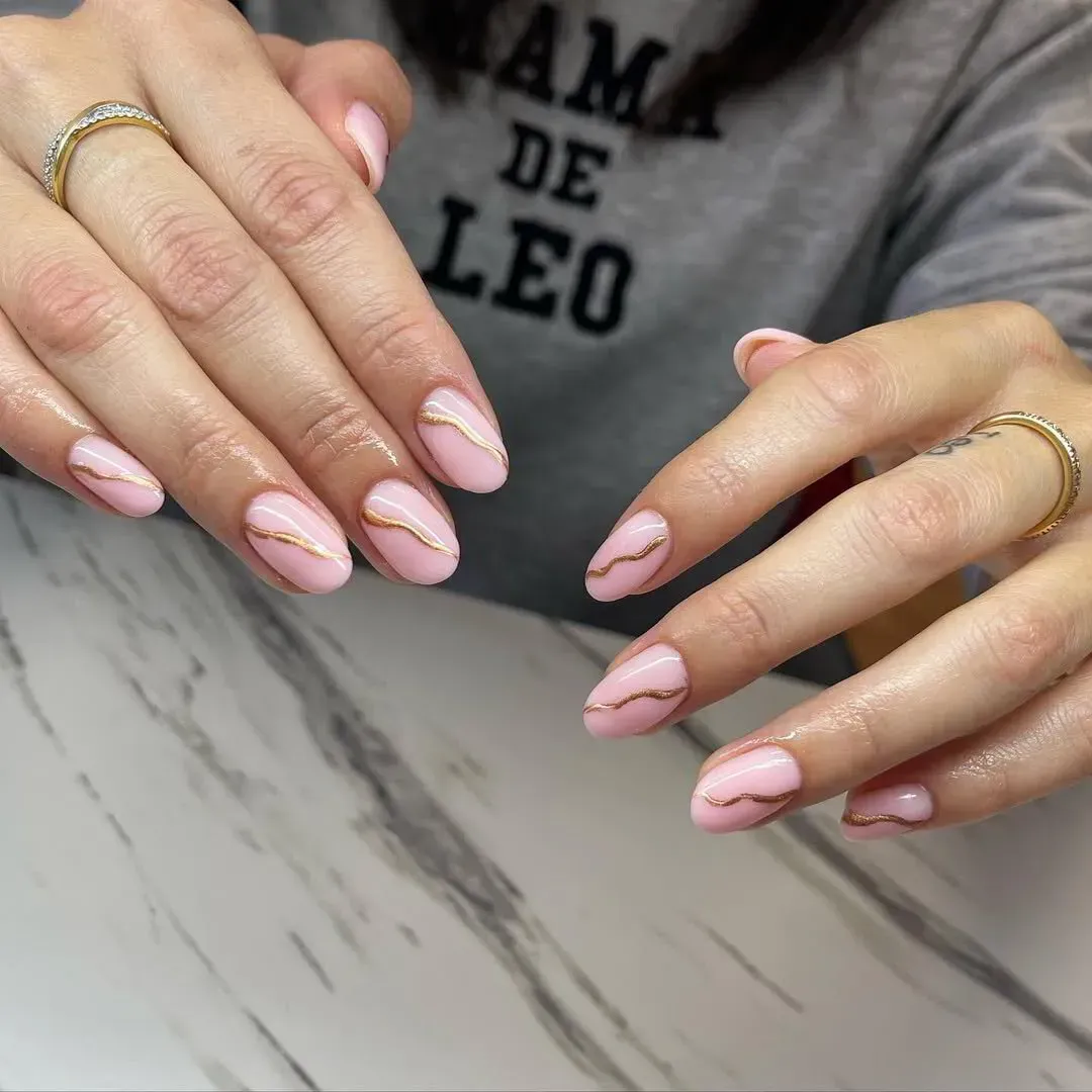 Uñas de color rosa claro con forma de almendra y una fina línea dorada ondulada