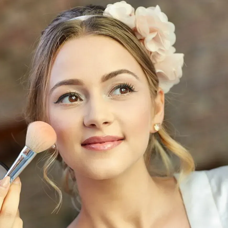 Una persona con un delicado adorno floral en el cabello se aplica colorete en las mejillas con una brocha de maquillaje.