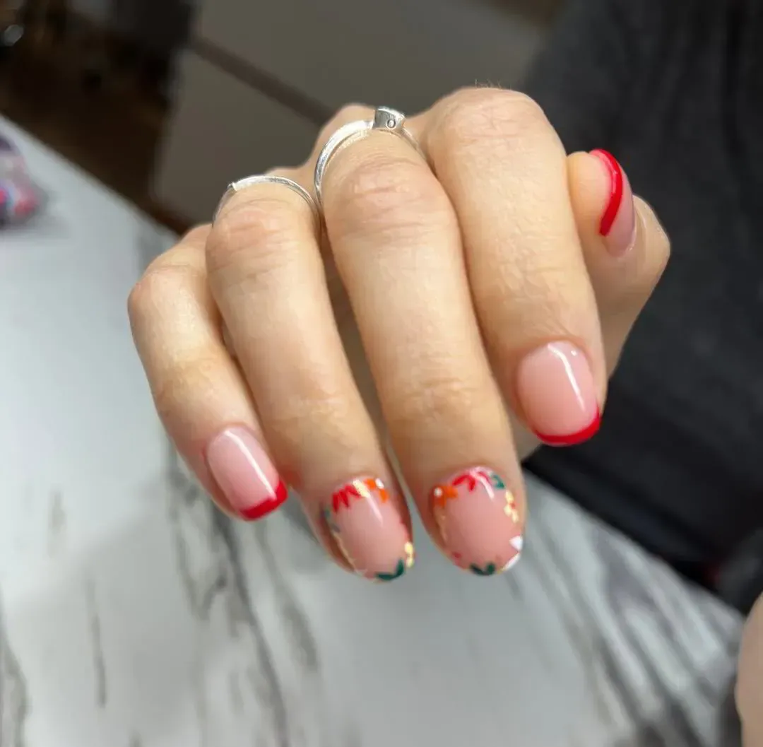 Una mano con manicura francesa en puntas rojas y coloridos detalles florales en los dedos medio y anular.