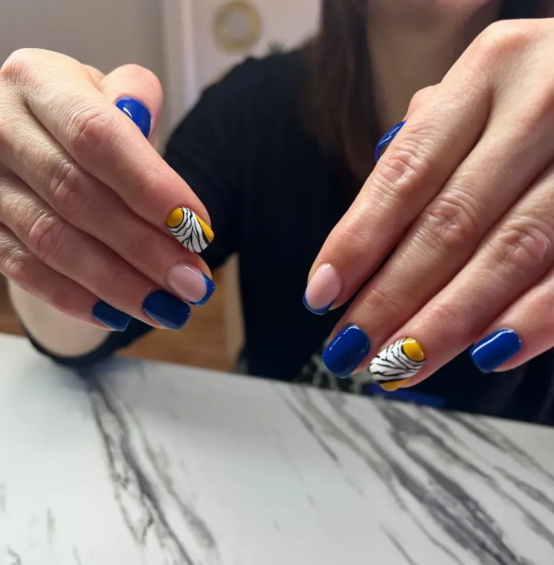 Manos con uñas bien cuidadas, con esmalte azul sólido, punta francesa y uñas con detalles amarillos y líneas blancas.