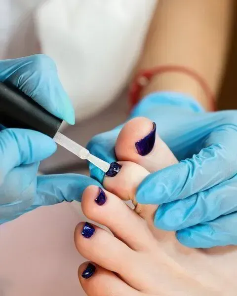 Una persona que lleva guantes azules aplica esmalte de uñas de color morado oscuro en una uña del pie durante una pedicura.