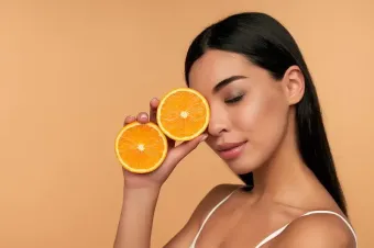 Una persona sostiene dos rodajas de naranja frente a un ojo sobre un fondo liso de tonos cálidos.