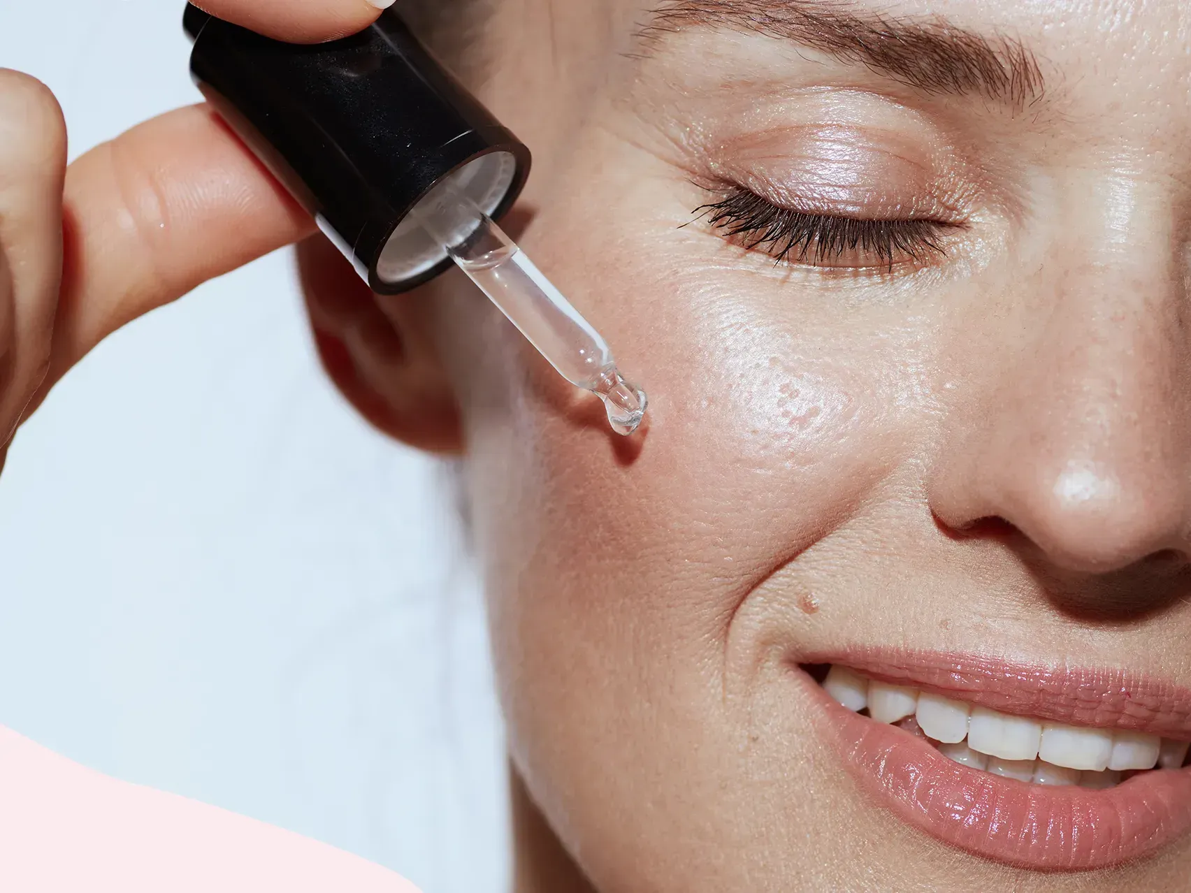 Una persona sonríe mientras se aplica un sérum facial transparente en la mejilla con un gotero de cristal.