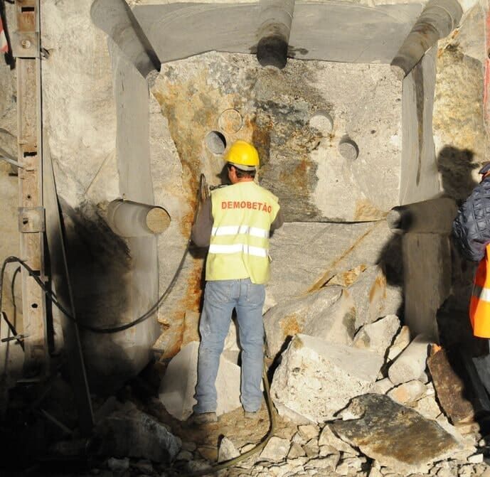 Trabalhador da construção civil perfurando uma parede de concreto, usando um colete amarelo e capacete.