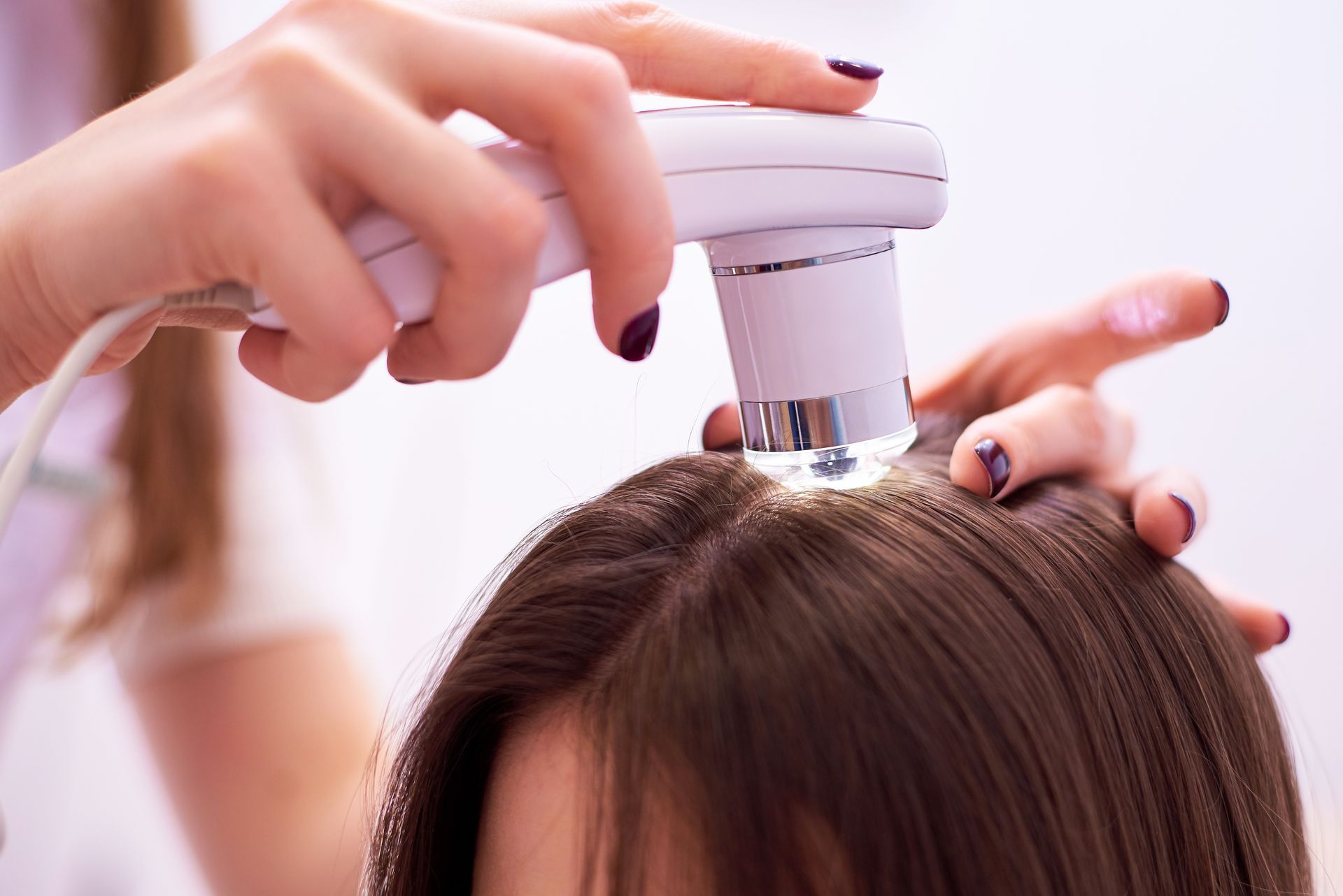 Instrument pour analyser le cuir chevelu au-dessus de cheveux bruns avec des mains vernies qui tiennent l'appareil et la tête de la personne.