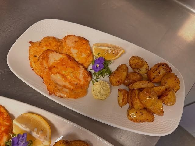 Ein weisser Teller mit gebratenem Fisch, Bratkartoffeln, einer Zitronenscheibe, einem Klecks Kräuterbutter und einer kleinen lila Blüte.