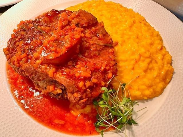 Eine Portion geschmortes Ossobuco mit roter Sauce, serviert zusammen mit leuchtend gelbem Safranrisotto auf einem weissen Teller.