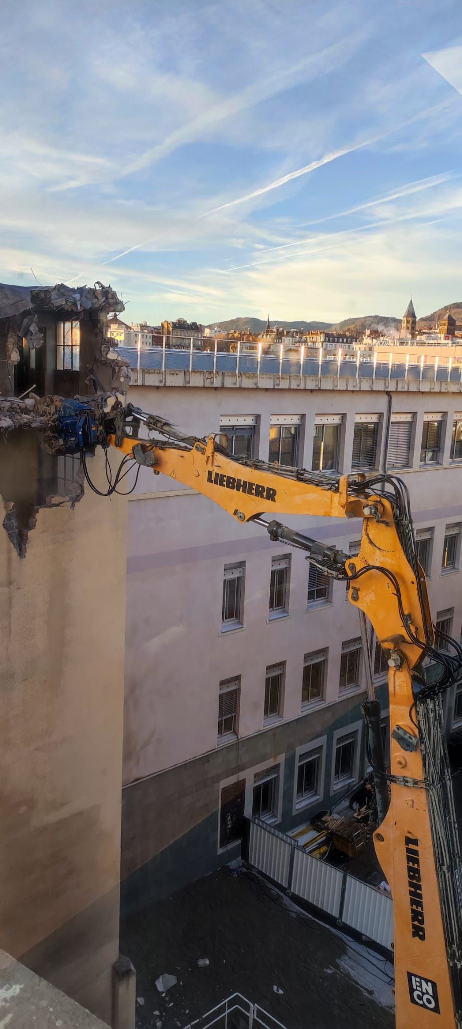 Une excavatrice démolit un bâtiment, le godet déchirant le toit contre un ciel bleu.