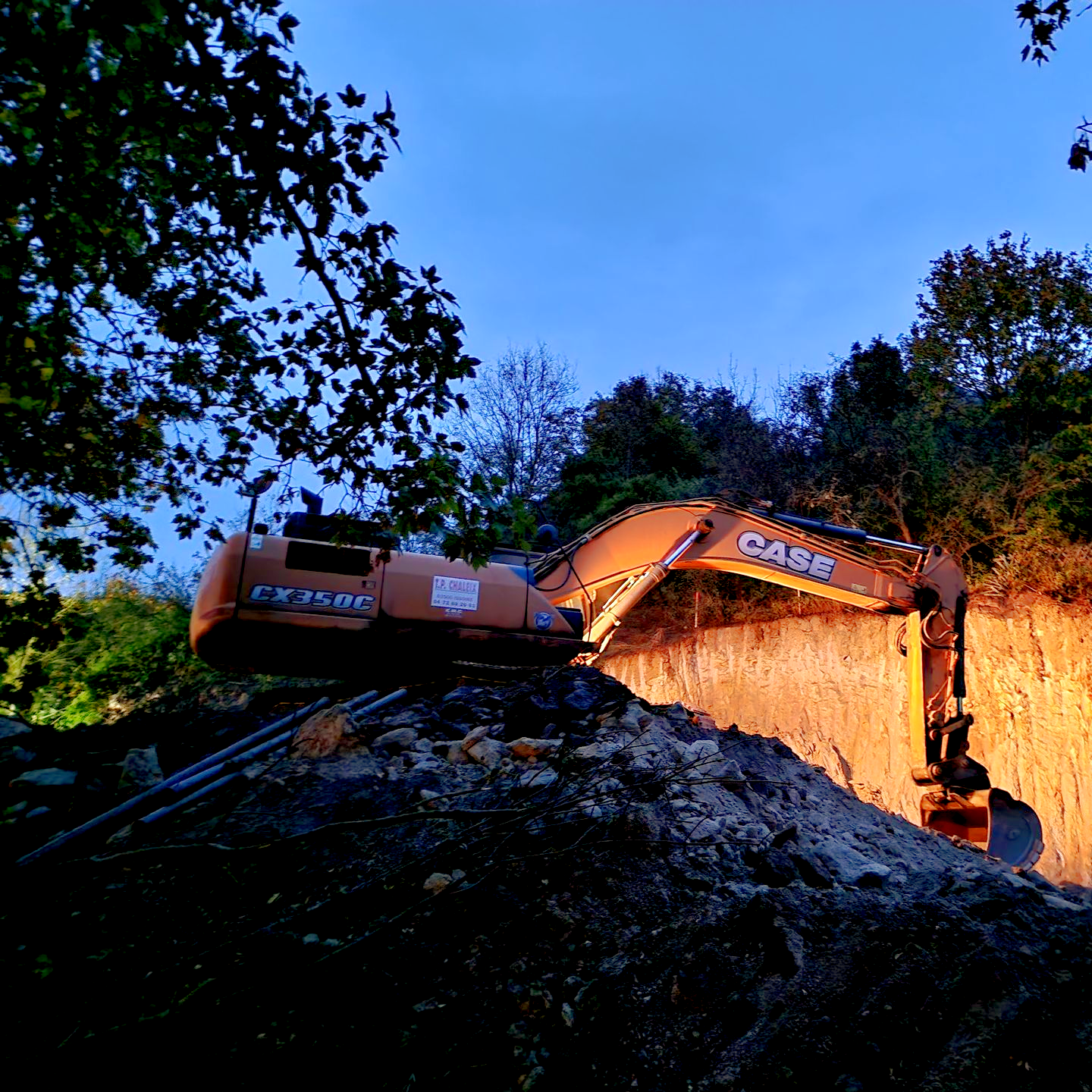 Excavatrice creusant un mur de terre au crépuscule. Machine orange et noire, éclairée par ses phares de travail.