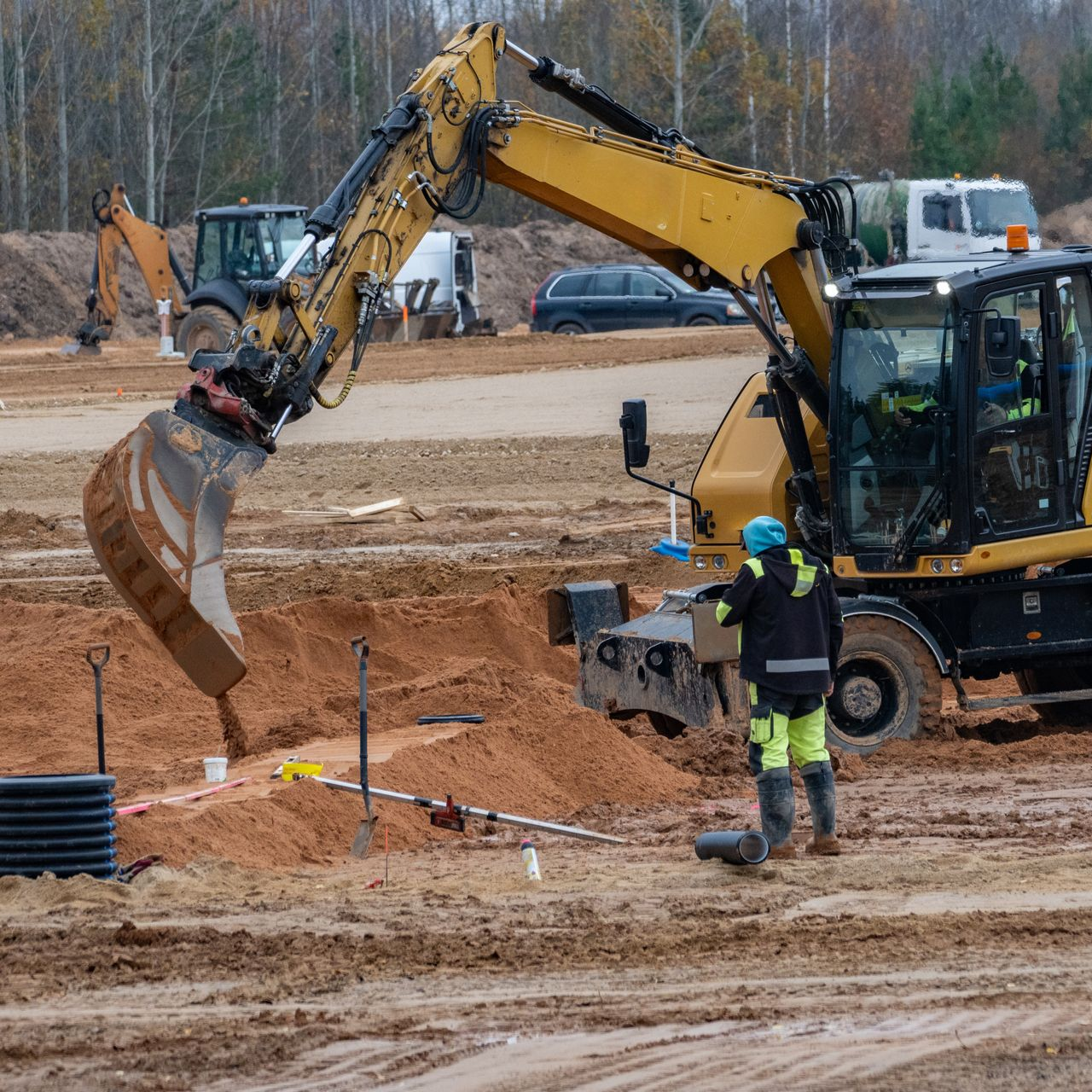 Une excavatrice jaune ramasse de la terre tandis qu'un ouvrier en tenue de haute visibilité observe sur un chantier de construction.