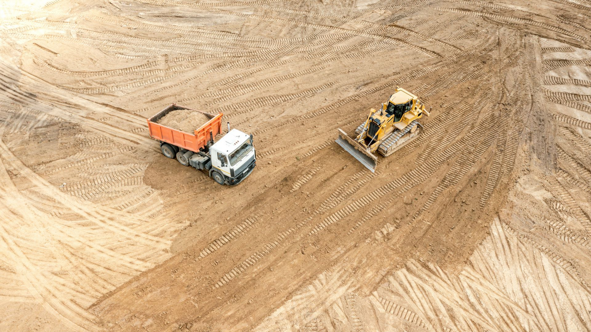 Chantier de construction avec un camion à benne basculante et un bulldozer travaillant sur une surface plane et sablonneuse.