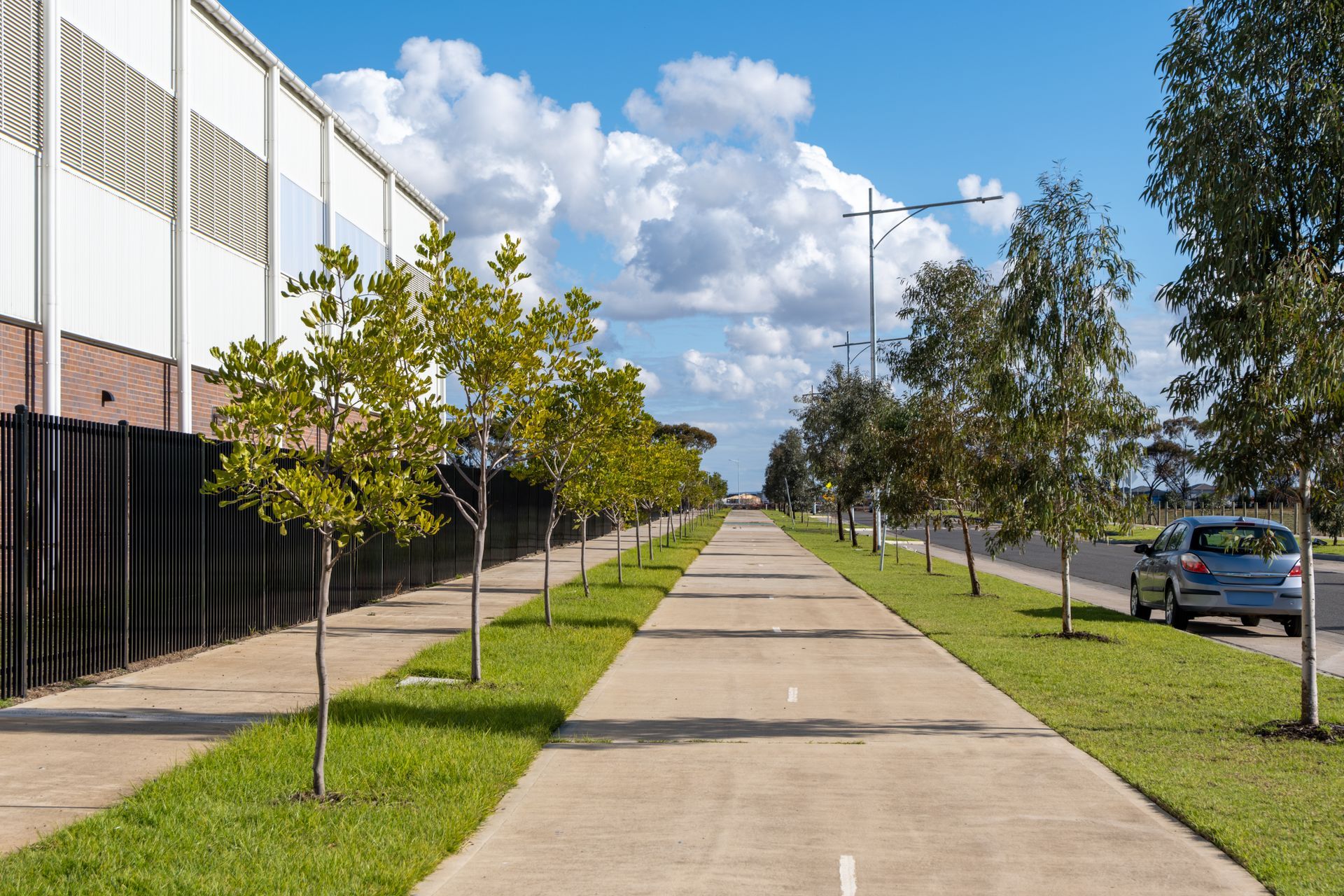 Chemin en béton bordé d'arbres et d'herbe, à côté d'un bâtiment et d'une clôture, sous un ciel partiellement nuageux.