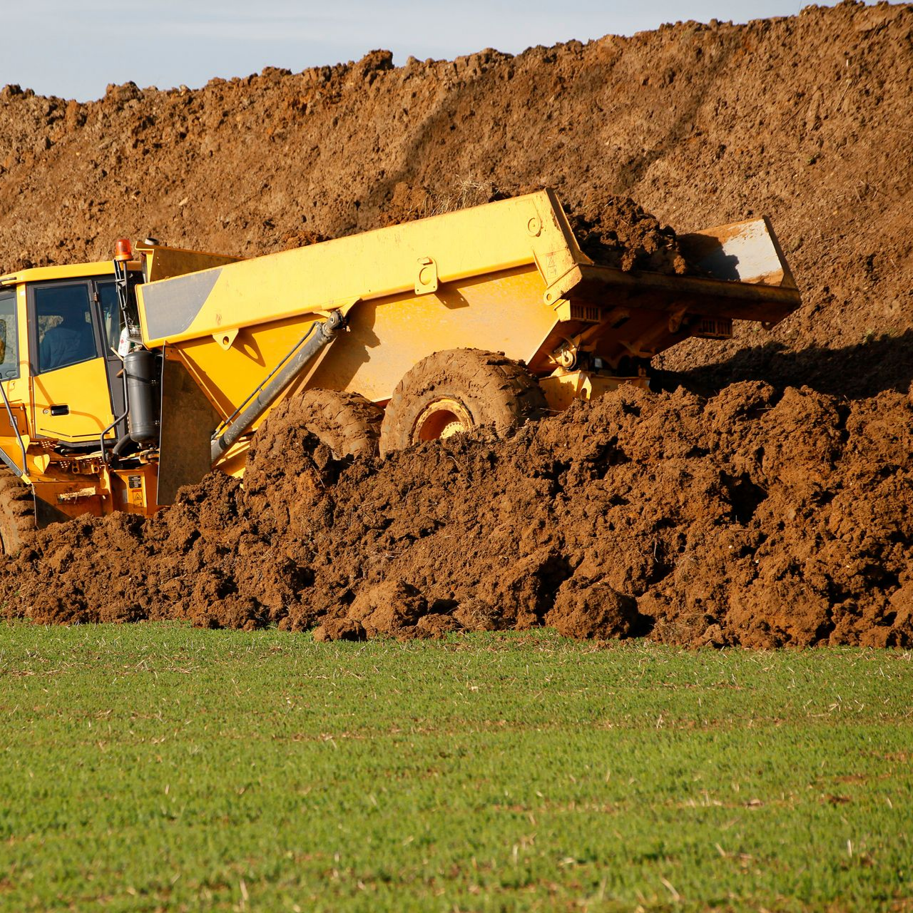 Camion à benne jaune coincé dans la boue près d'un tas de terre, champ au premier plan.