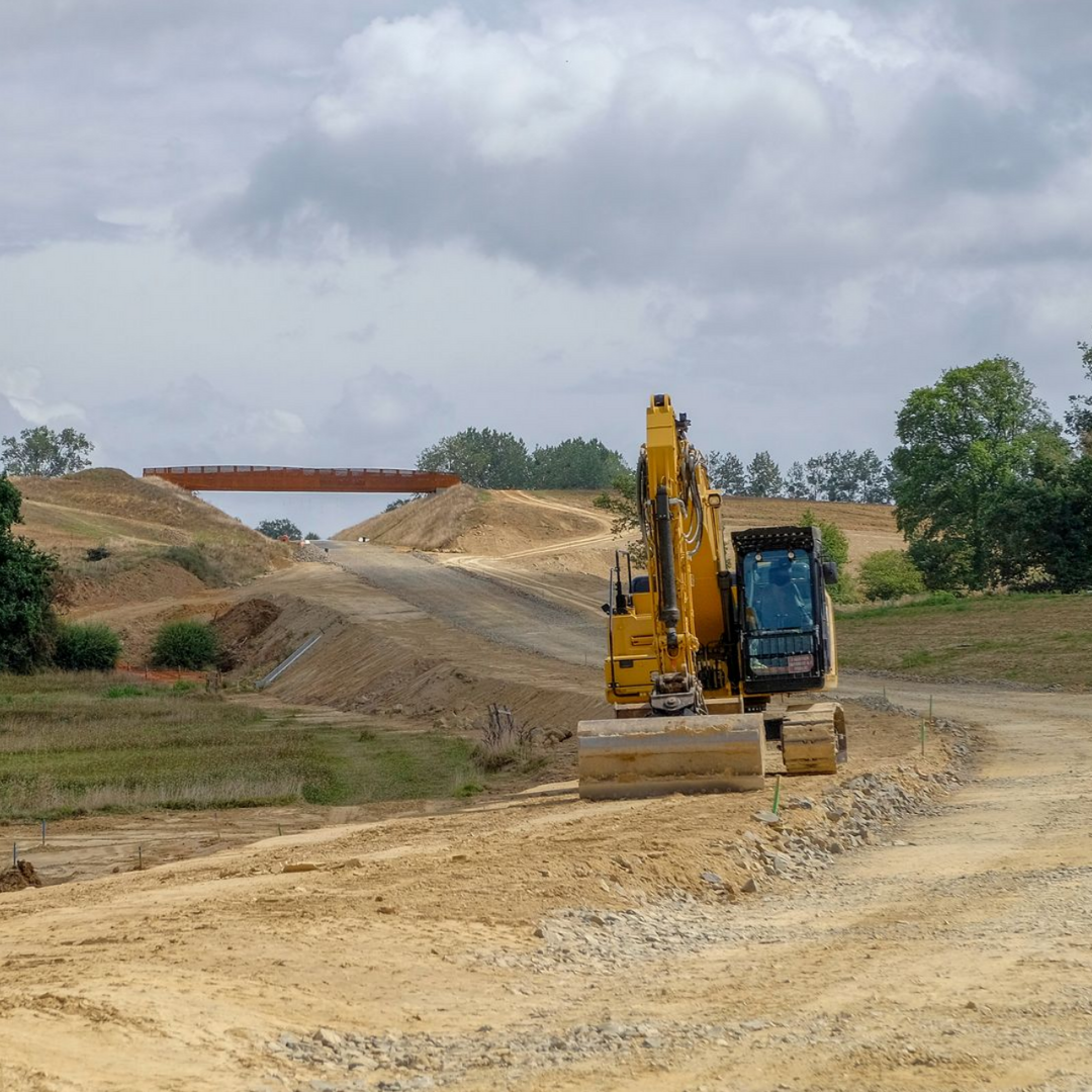 Excavatrice jaune sur un chemin de terre, nivelant la terre, avec un pont au loin sous un ciel nuageux.