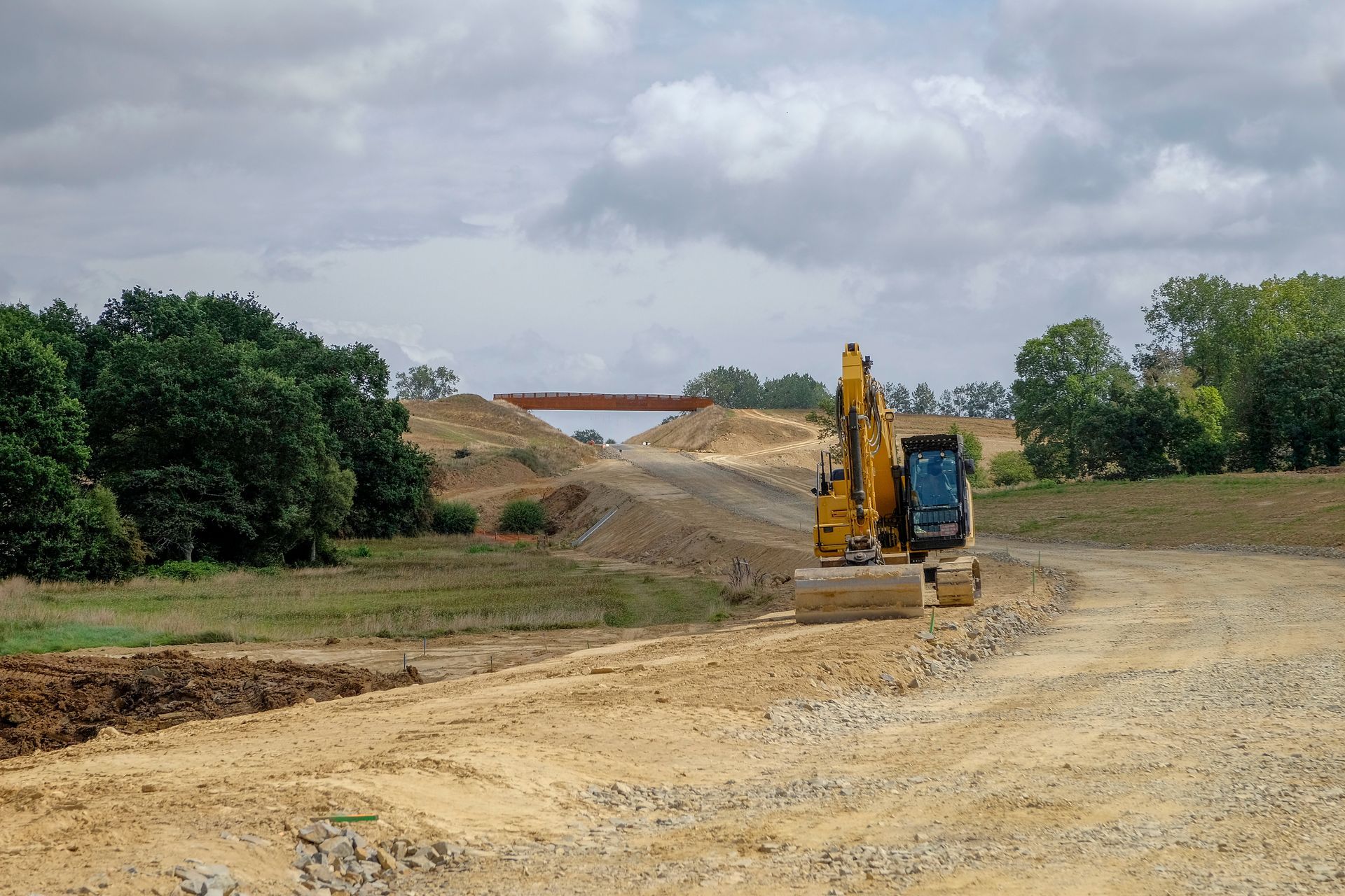 Chantier de construction avec une excavatrice jaune, des collines de terre et un ciel nuageux.
