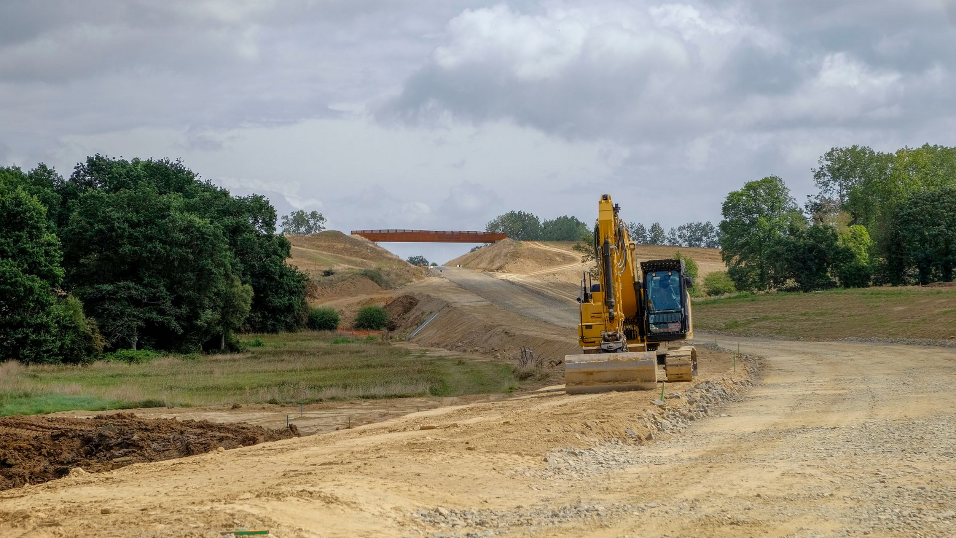Chantier de construction avec excavatrice jaune, terrassement et pont sous un ciel nuageux.