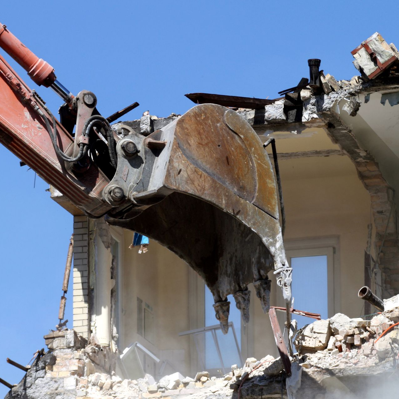 Une excavatrice démolit un bâtiment, le godet déchirant le toit contre un ciel bleu.