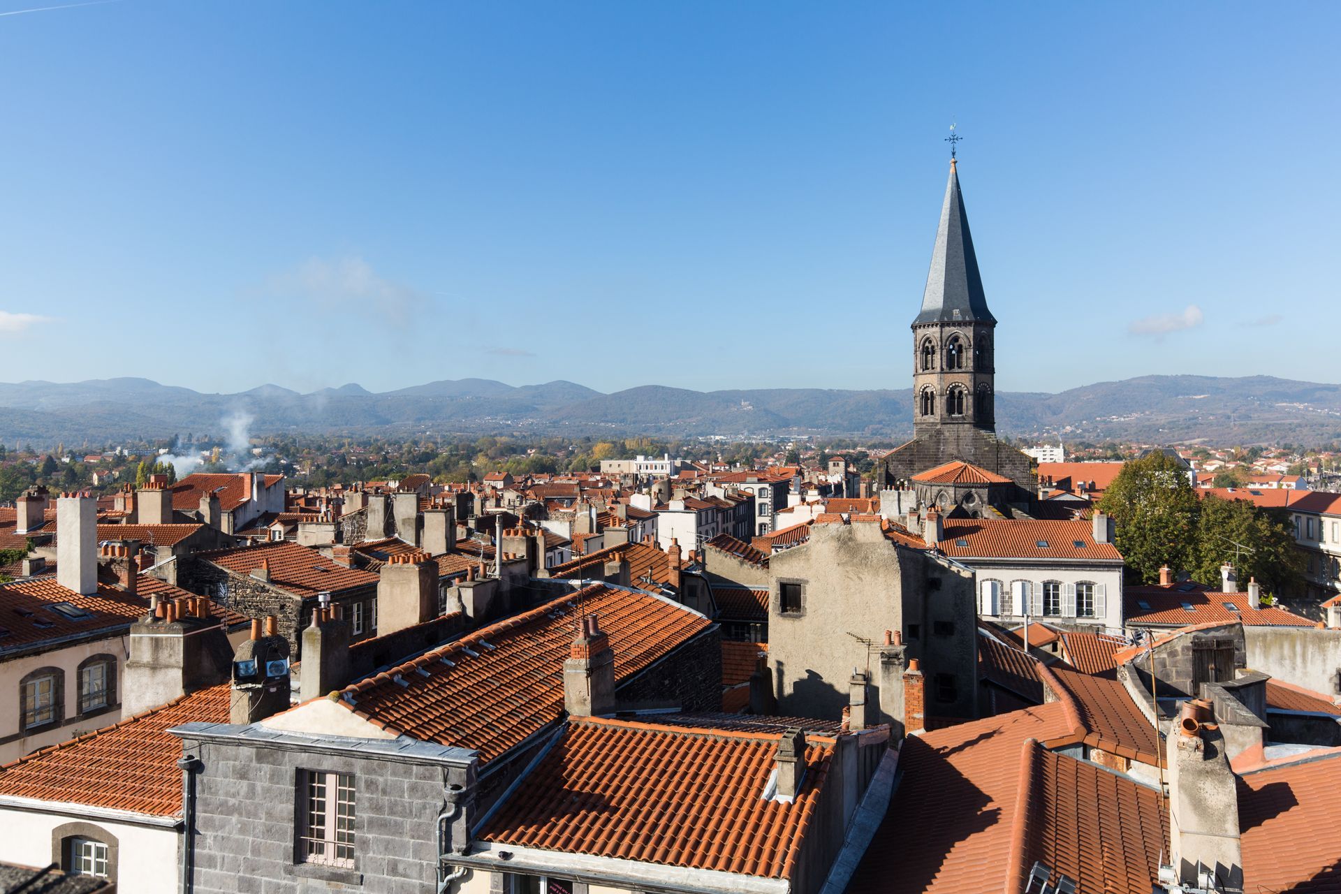 Paysage urbain avec des toits de tuiles orange, des bâtiments en pierre et un grand clocher sous un ciel bleu clair.
