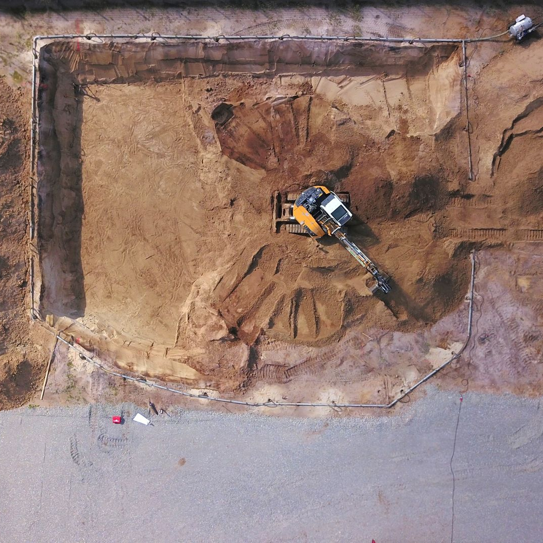 Vue aérienne d'une excavatrice creusant une fosse de fondation rectangulaire dans un terrain en terre.
