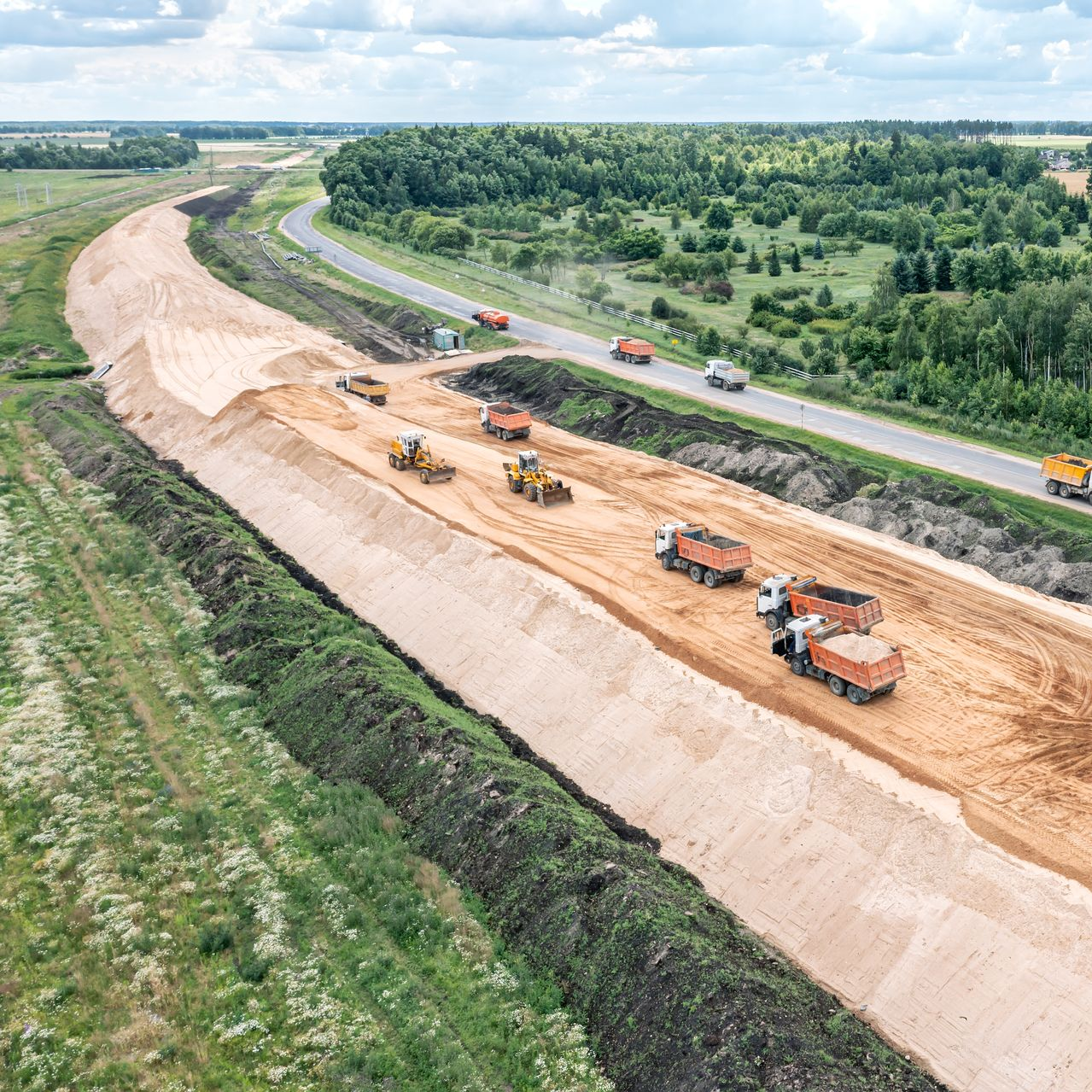 Chantier de construction avec machinerie lourde, camions à benne basculante et autoroute.