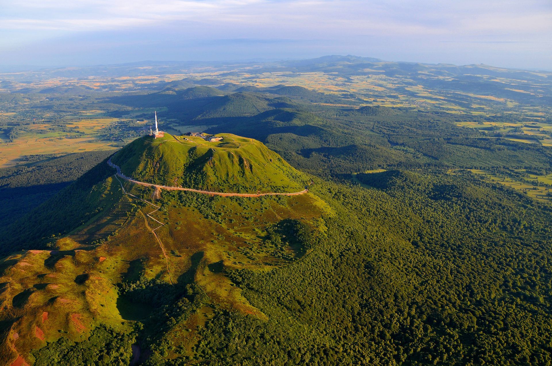 Vue aérienne d'une montagne volcanique verte avec une tour de communication, entourée de forêts et d'un paysage lointain.