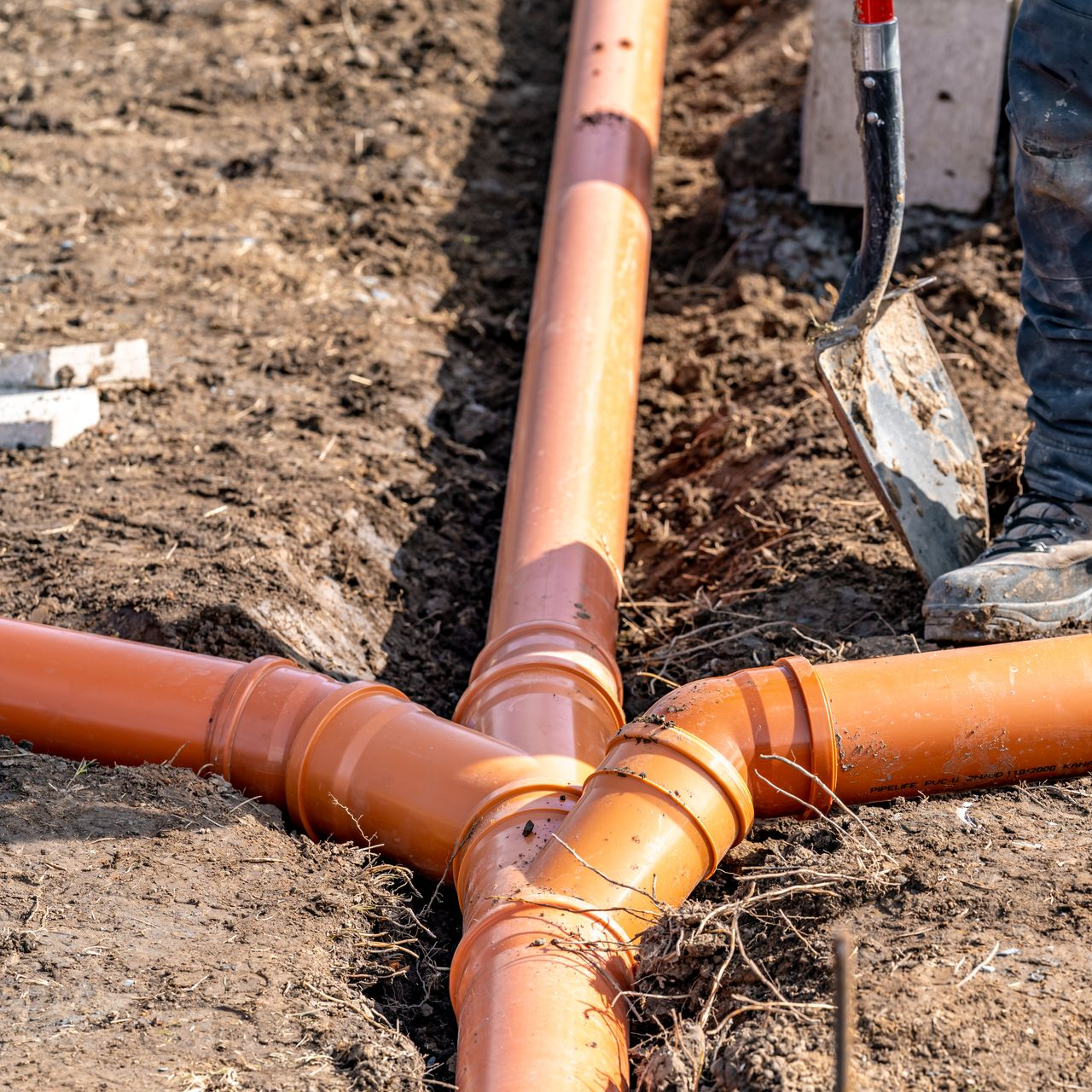 Des tuyaux de drainage orange sont installés dans une tranchée ; un ouvrier avec une pelle se tient à proximité.