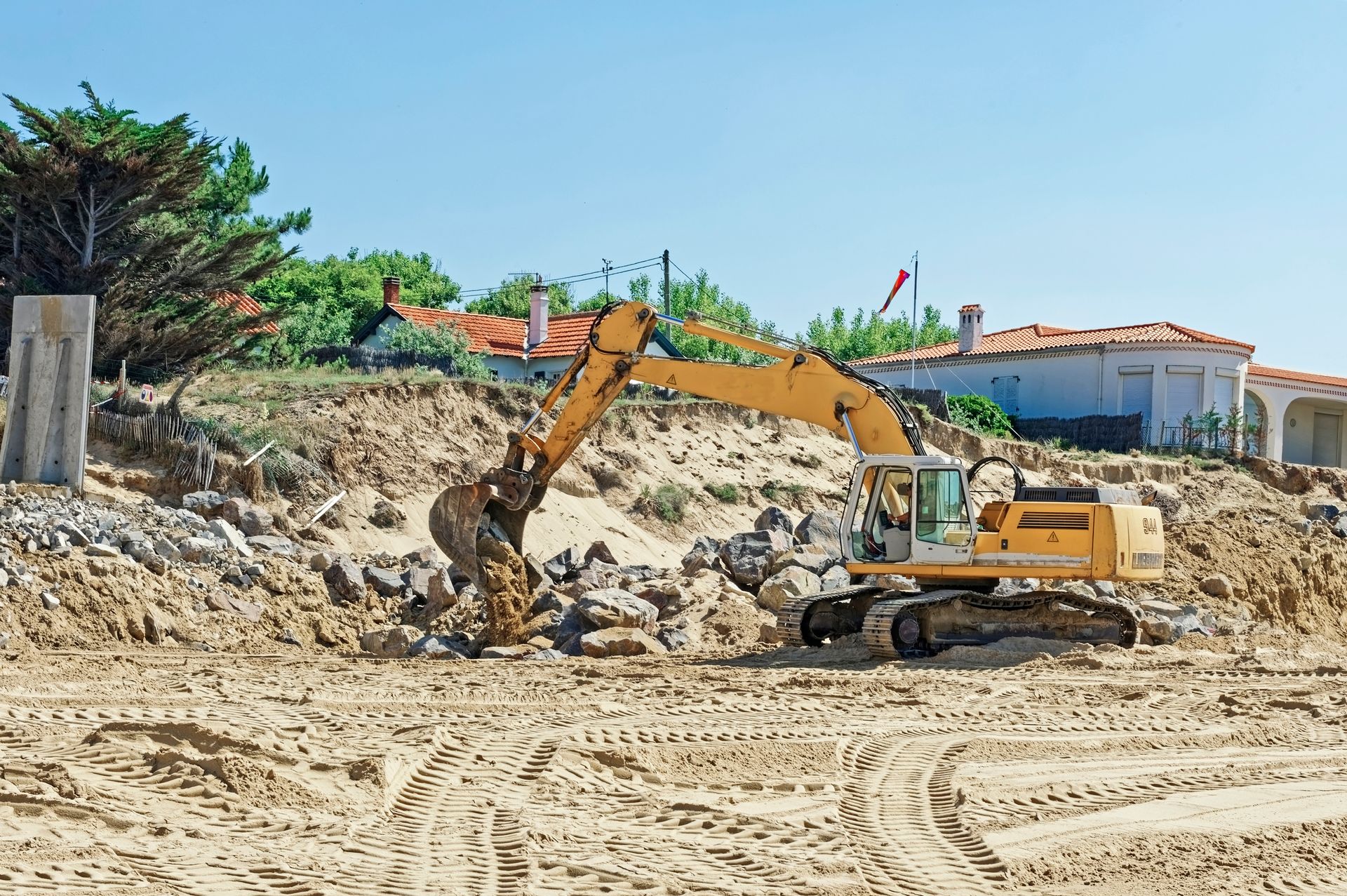Excavatrice jaune sur un sol sablonneux, près de rochers, de maisons et d'arbres sous un ciel bleu clair.