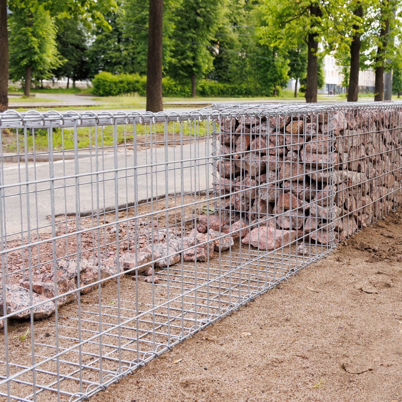 Mur de gabions en treillis métallique, garni de pierres rougeâtres. Extérieur, sol en terre, arbres en arrière-plan.