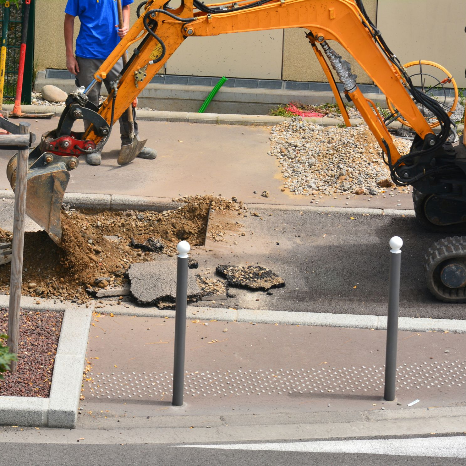 Une excavatrice creuse le trottoir, à côté de bornes et d'un ouvrier en chemise bleue.