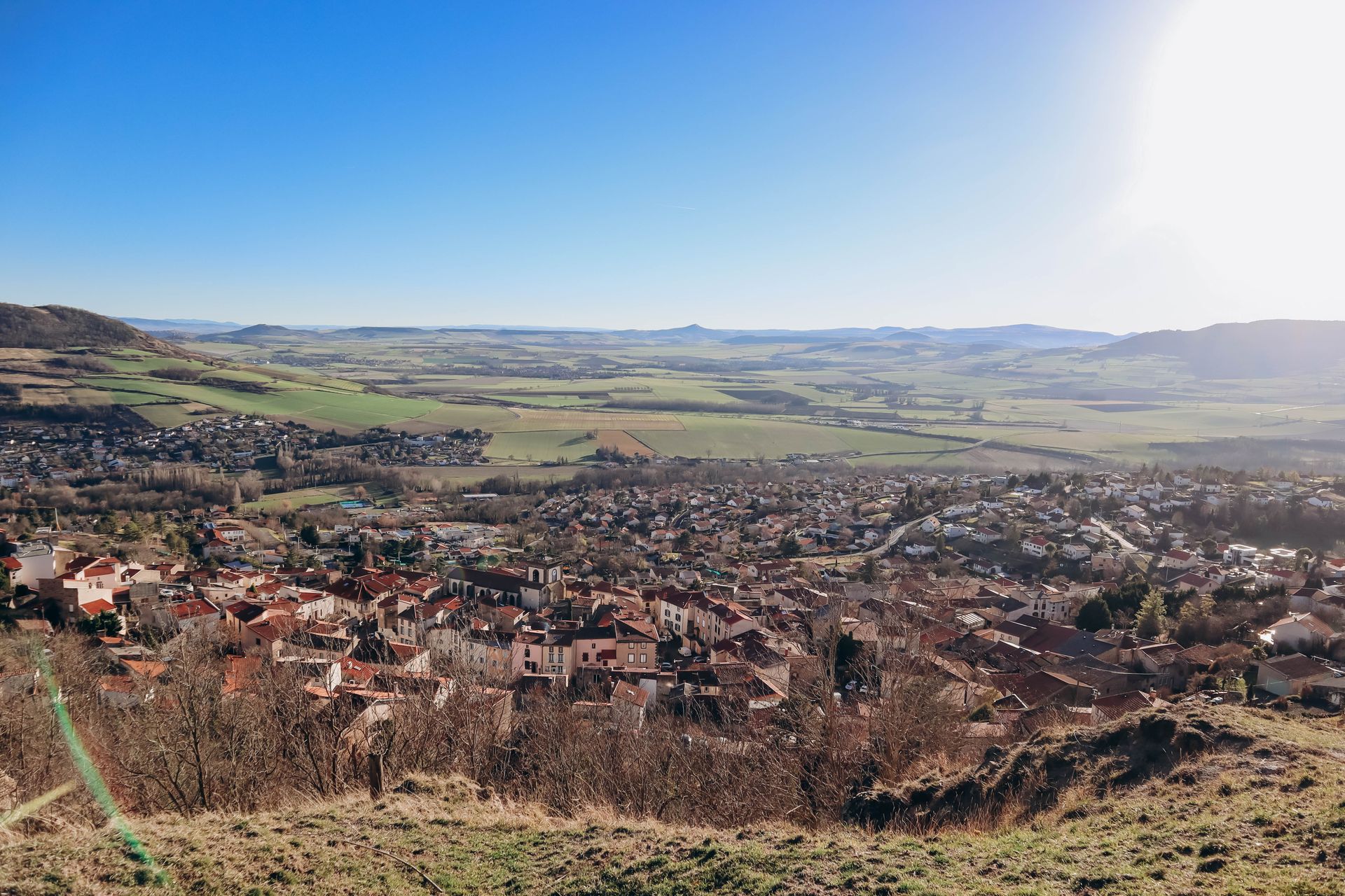 Vue panoramique d'une ville nichée dans une vallée, avec des champs verdoyants et des collines sous un ciel bleu vif.