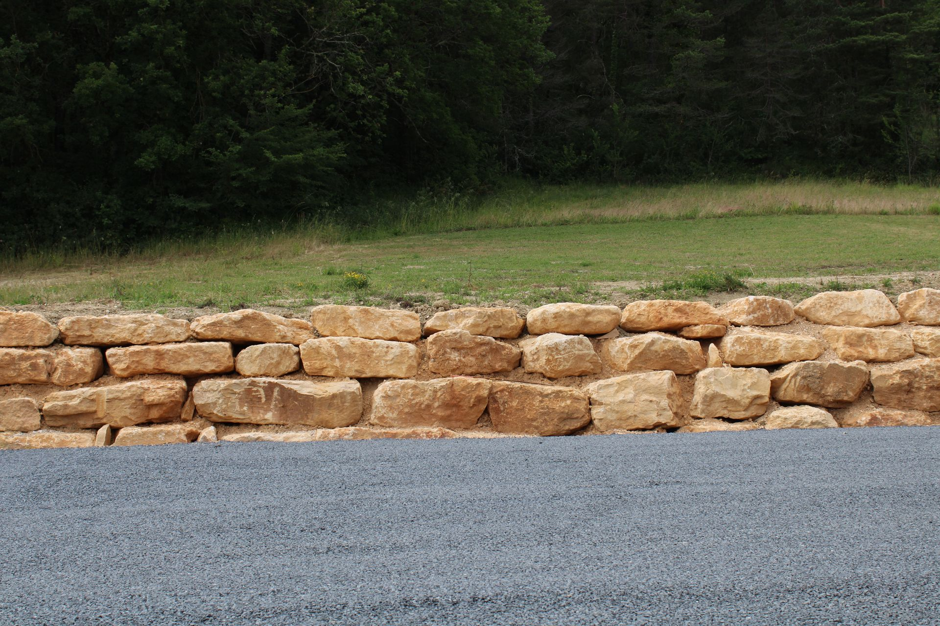 Mur de soutènement en pierre avec une base en gravier devant une colline herbeuse.