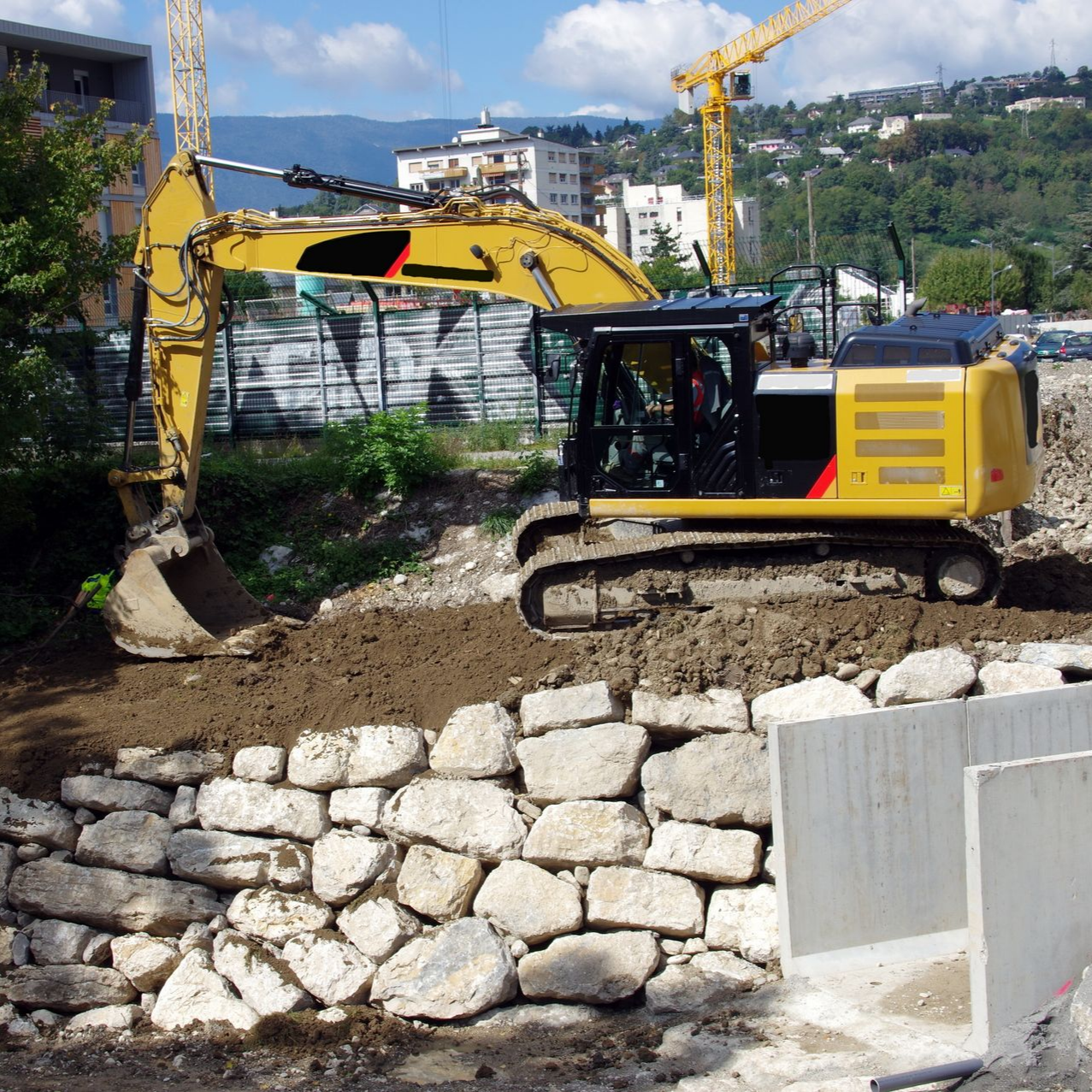 Une excavatrice jaune creuse sur un chantier près d'un mur de pierre. Une grue est visible à l'arrière-plan.