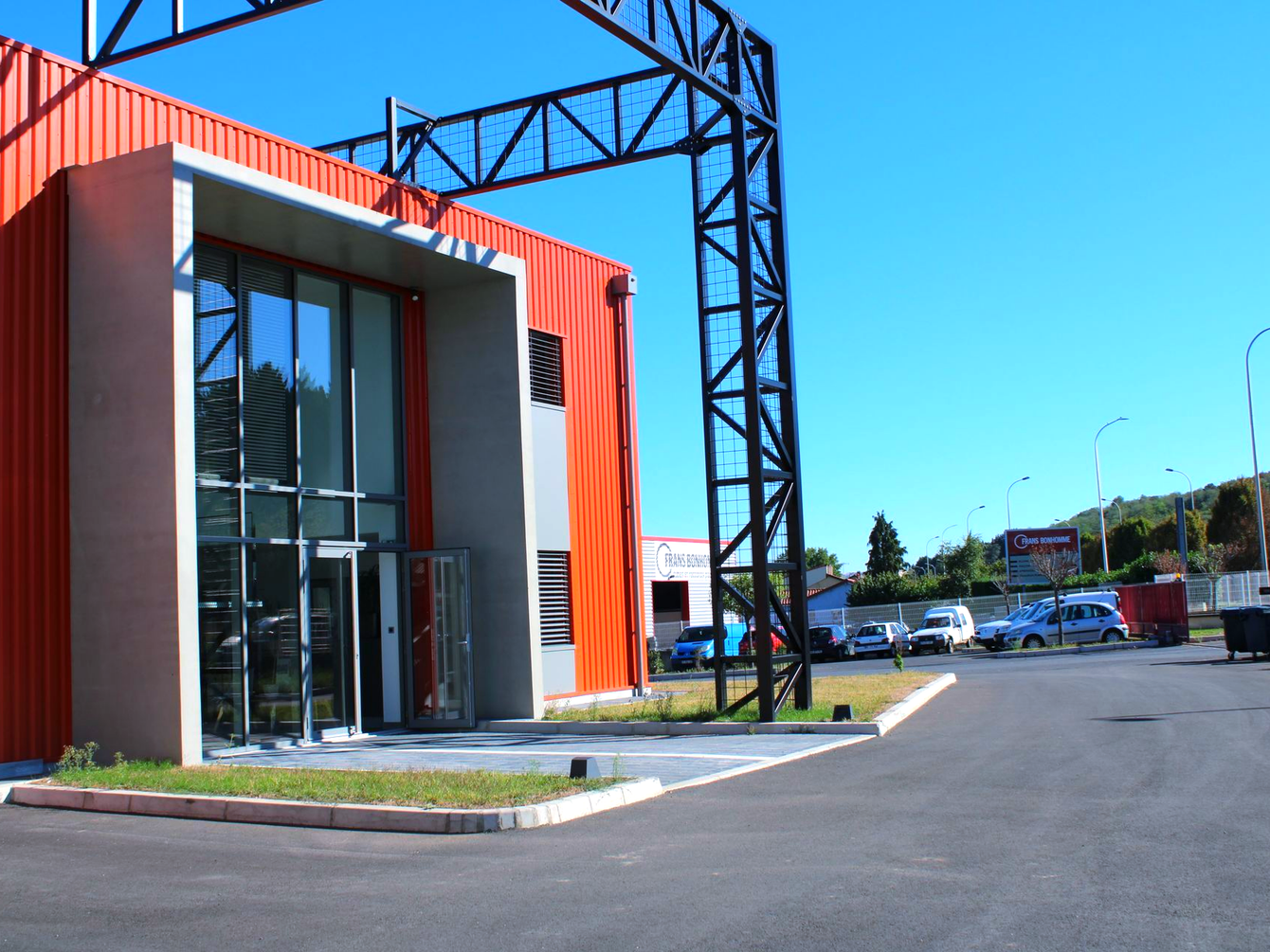 Bâtiment industriel rouge et gris avec portes vitrées, poutres métalliques noires et parking.