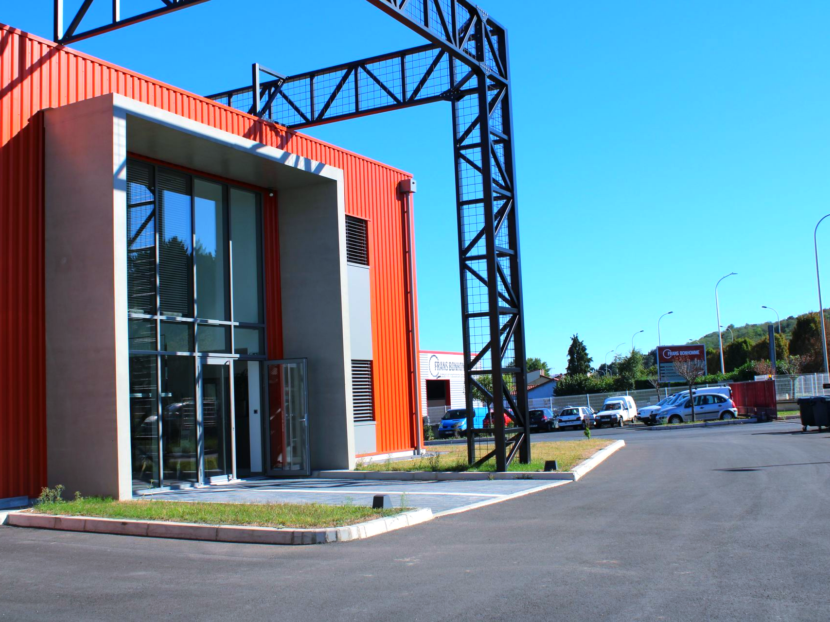 Bâtiment industriel rouge et gris avec portes vitrées, poutres métalliques noires et parking.