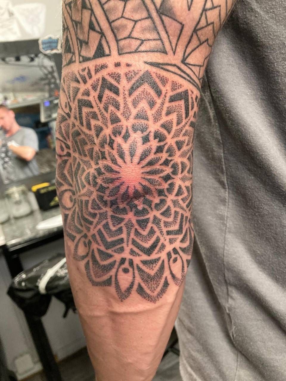 Ein schwarz-graues geometrisches Mandala-Tattoo, zentriert auf dem Ellbogen, mit zusätzlichen Mustern im Stammesstil am Oberarm.