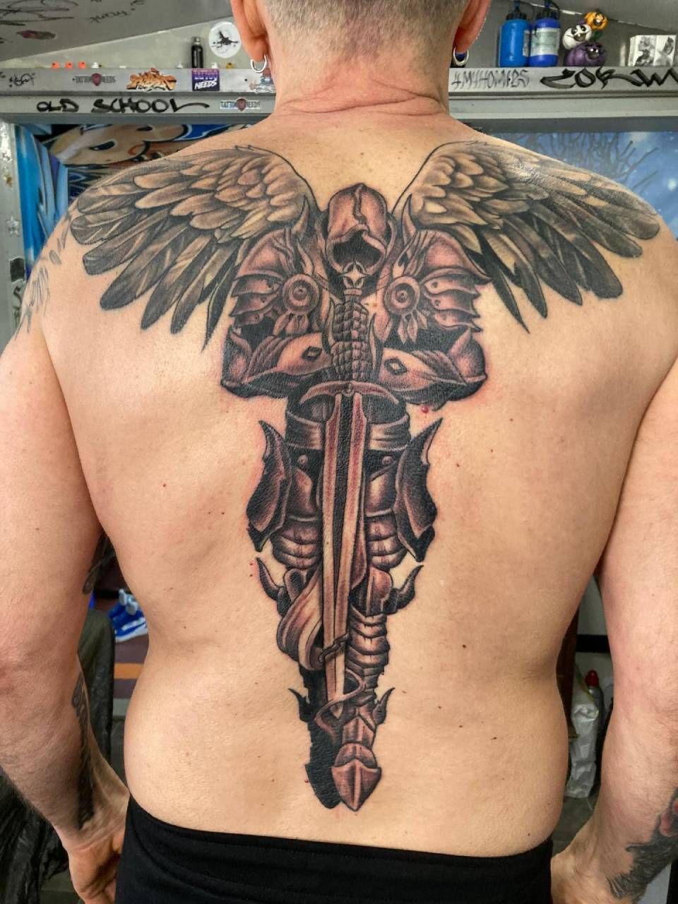 Ein schwarz-graues Rückentattoo, das einen geflügelten, gepanzerten Ritter zeigt, der ein grosses Schwert umklammert, das sich die Wirbelsäule hinunter erstreckt.