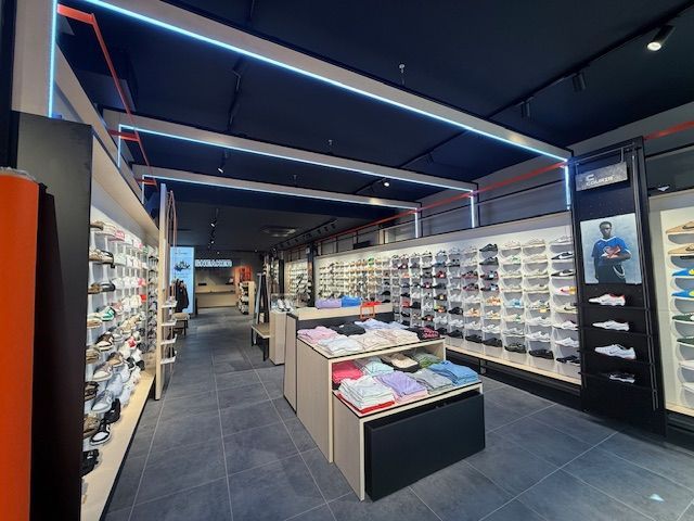 L'intérieur d'un magasin de chaussures de sport bien éclairé, avec des rangées de baskets sur des présentoirs muraux et une table pliante centrale présentant des vêtements.