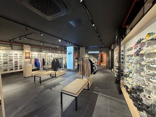 L'intérieur d'un magasin de chaussures présentant des rangées de baskets sur des présentoirs muraux, des portants à vêtements et des bancs sur un sol carrelé foncé.