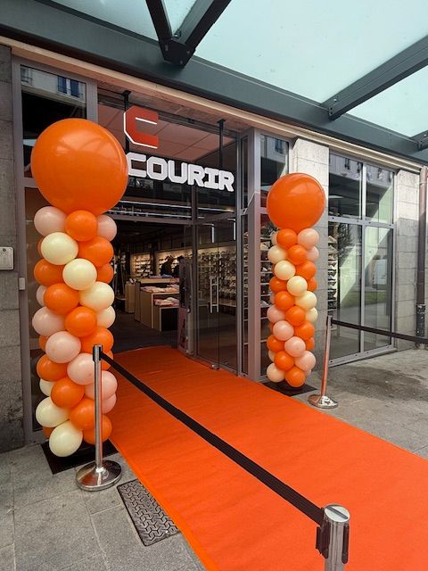 L'entrée d'un magasin, recouverte d'un tapis orange, est flanquée de deux colonnes de ballons surmontées de gros ballons orange.