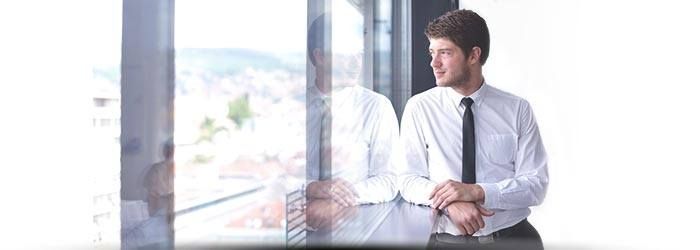 Un homme en chemise blanche et cravate est assis à un bureau près d'une fenêtre lumineuse, regardant dehors.